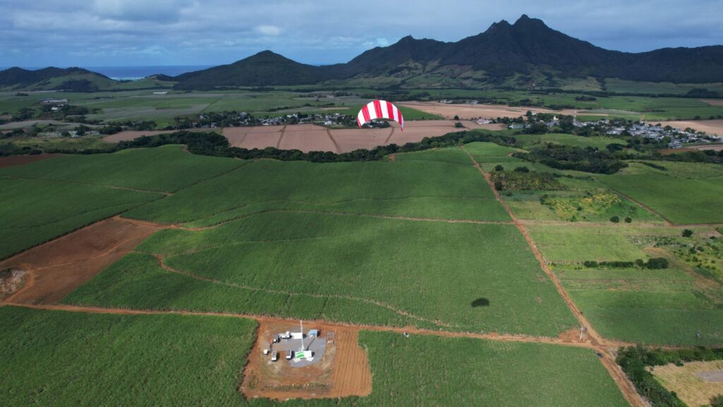 A SkySails airborne wind energy system featuring a kite in the power phase soaring above the green sugarcane fields of Mauritius, with mountains to the right and the ocean to the left.